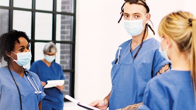 Foto von vier Pflegekräften in blauen Kitteln, mit medizinischen Masken und Klemmbrettern im Gespräch.