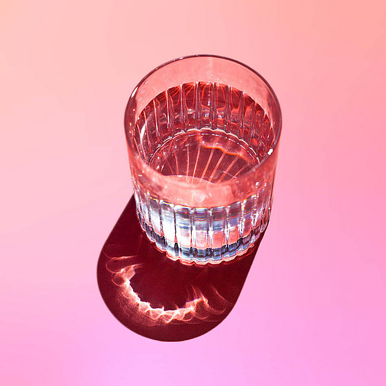 Ein klares Glas Wasser wirft einen bunten Schatten auf einen rosa-orangefarbenen Hintergrund mit Farbverlauf. Das Licht erzeugt durch das Glas und den Schatten komplizierte Muster und hebt das Farbenspiel hervor.