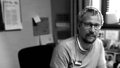 Schwarz-Weiß-Porträt von Andreas, der an einem Schreibtisch sitzt und eine Brille trägt.